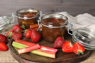 Photo of Jars of tasty rhubarb jam, fresh stems and strawberries on wooden table