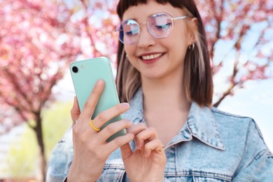 Photo of Beautiful young woman with phone near blossoming sakura tree in park, focus on hands