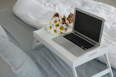 Photo of White tray table with laptop and bouquet of beautiful daisies on bed