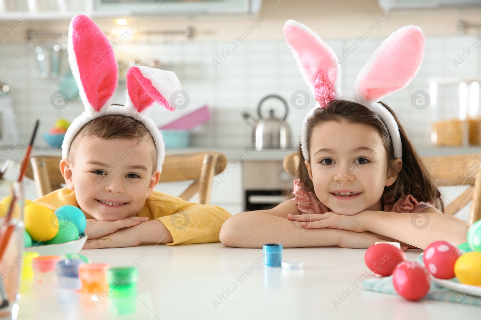 Photo of Cute children with bunny ears headbands and painted Easter eggs sitting at table in kitchen