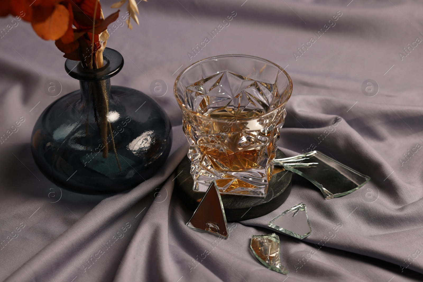 Photo of Glass of tasty alcohol drink, shards of broken mirror and vase with decorative flowers on grey fabric