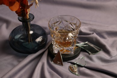 Glass of tasty alcohol drink, shards of broken mirror and vase with decorative flowers on grey fabric