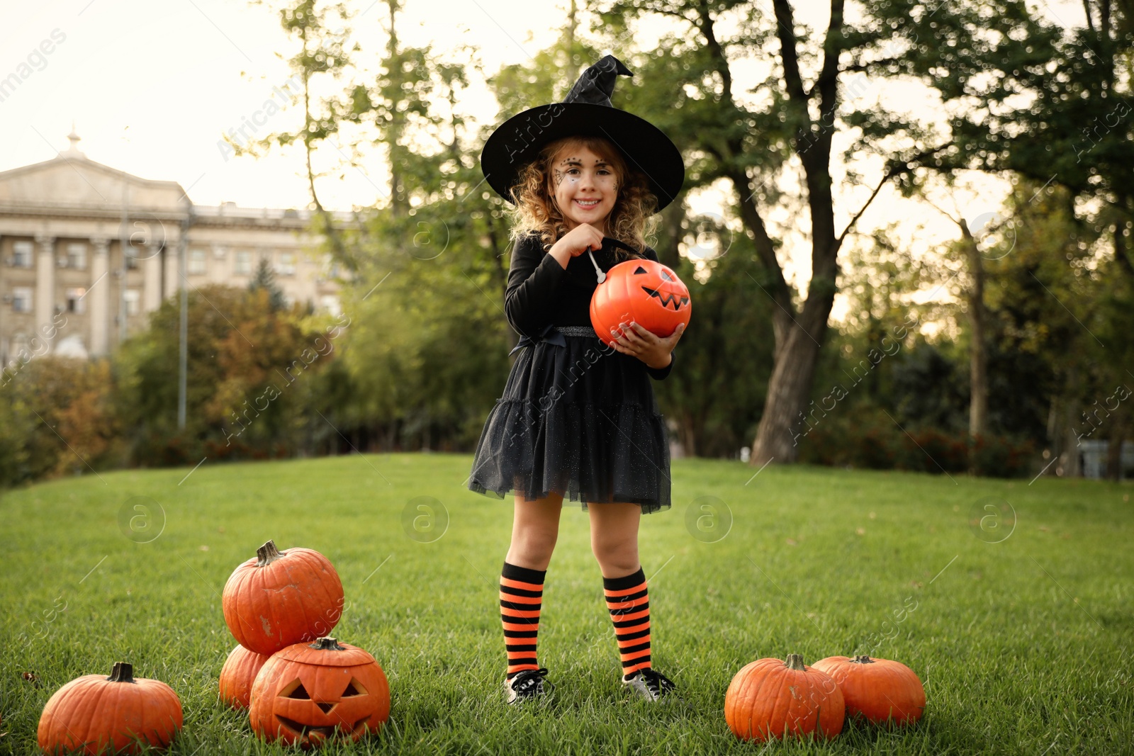 Photo of Cute little girl with pumpkin candy bucket wearing Halloween costume in park