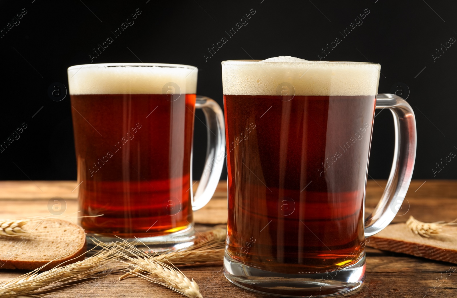 Photo of Mugs of delicious kvass, spikes and bread on wooden table against black background