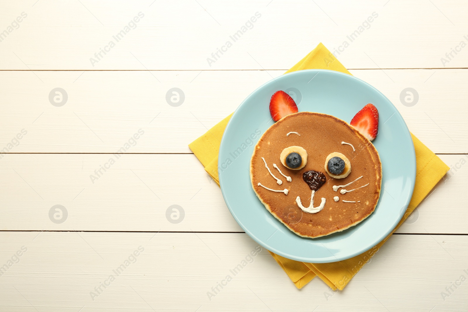 Photo of Creative serving for kids. Plate with cute cat made of pancakes, berries, cream, banana and chocolate paste on white wooden table, top view. Space for text