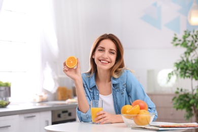 Photo of Happy young woman with glass of juice and orange at table in kitchen. Healthy diet