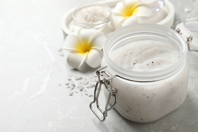 Photo of Body scrub in glass jar and plumeria flowers on grey marble table, closeup. Space for text