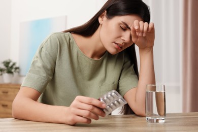 Sad woman with blister of pills suffering from headache at wooden table indoors