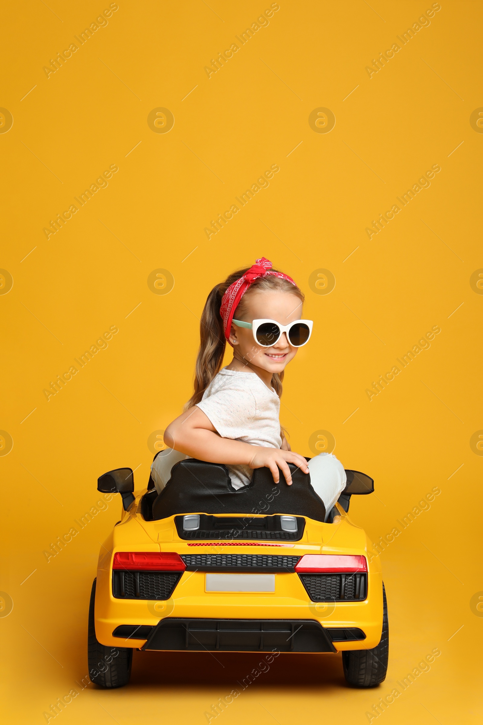 Photo of Cute little girl driving children's electric toy car on yellow background