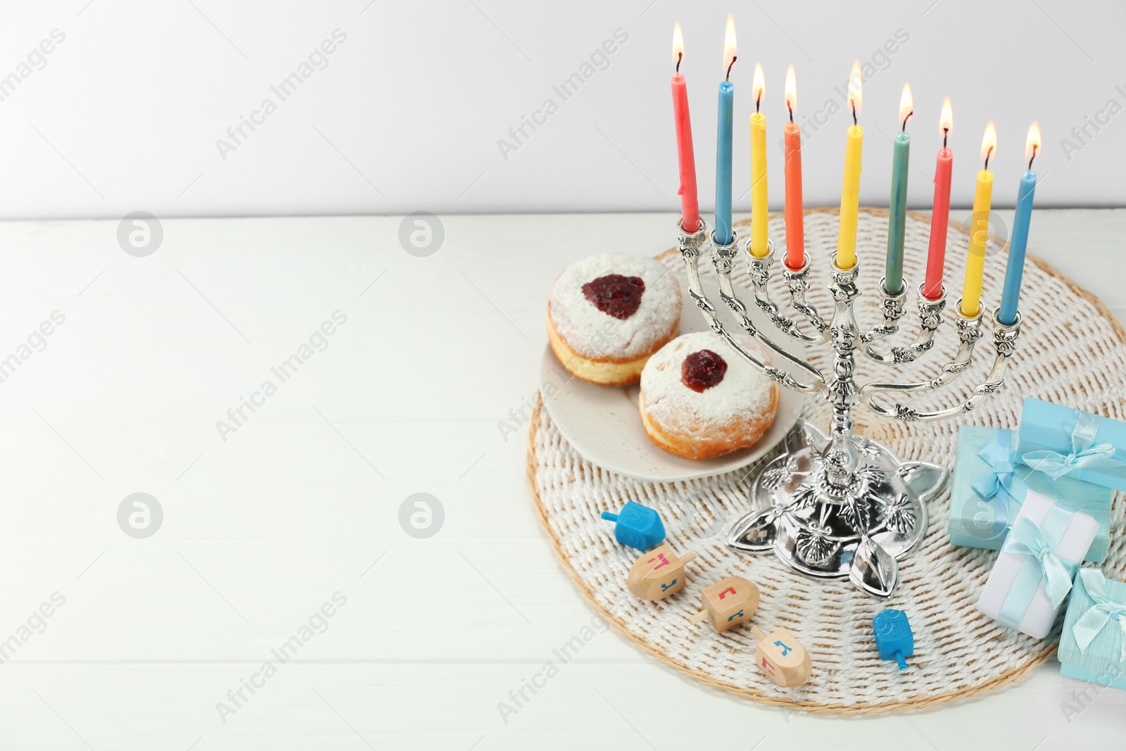 Photo of Hanukkah celebration. Menorah with burning candles, dreidels, donuts and gift boxes on white wooden table, space for text