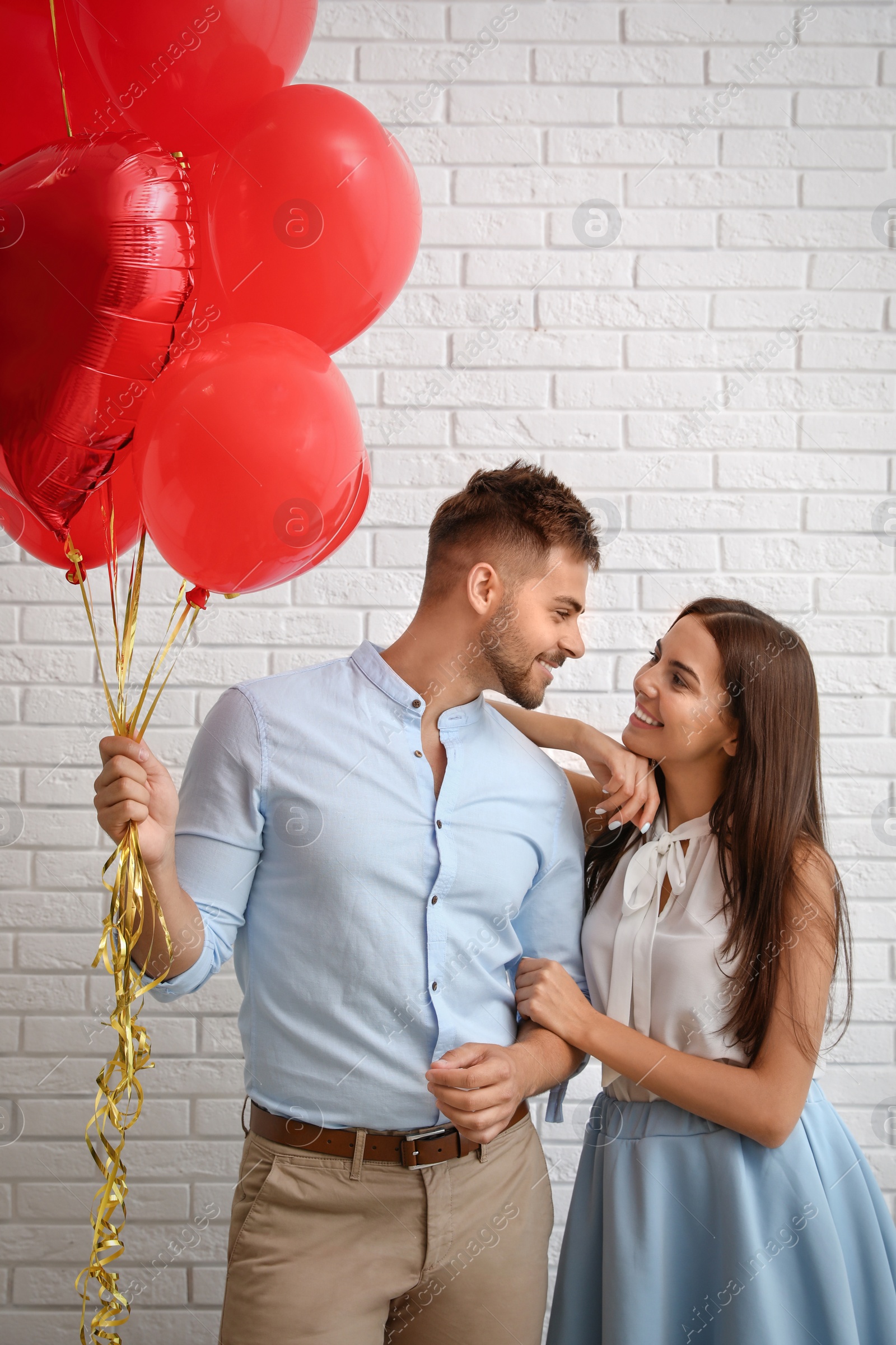 Photo of Young couple with air balloons near white brick wall. Celebration of Saint Valentine's Day