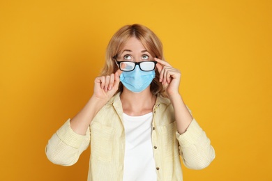 Woman wiping foggy glasses caused by wearing medical mask on yellow background