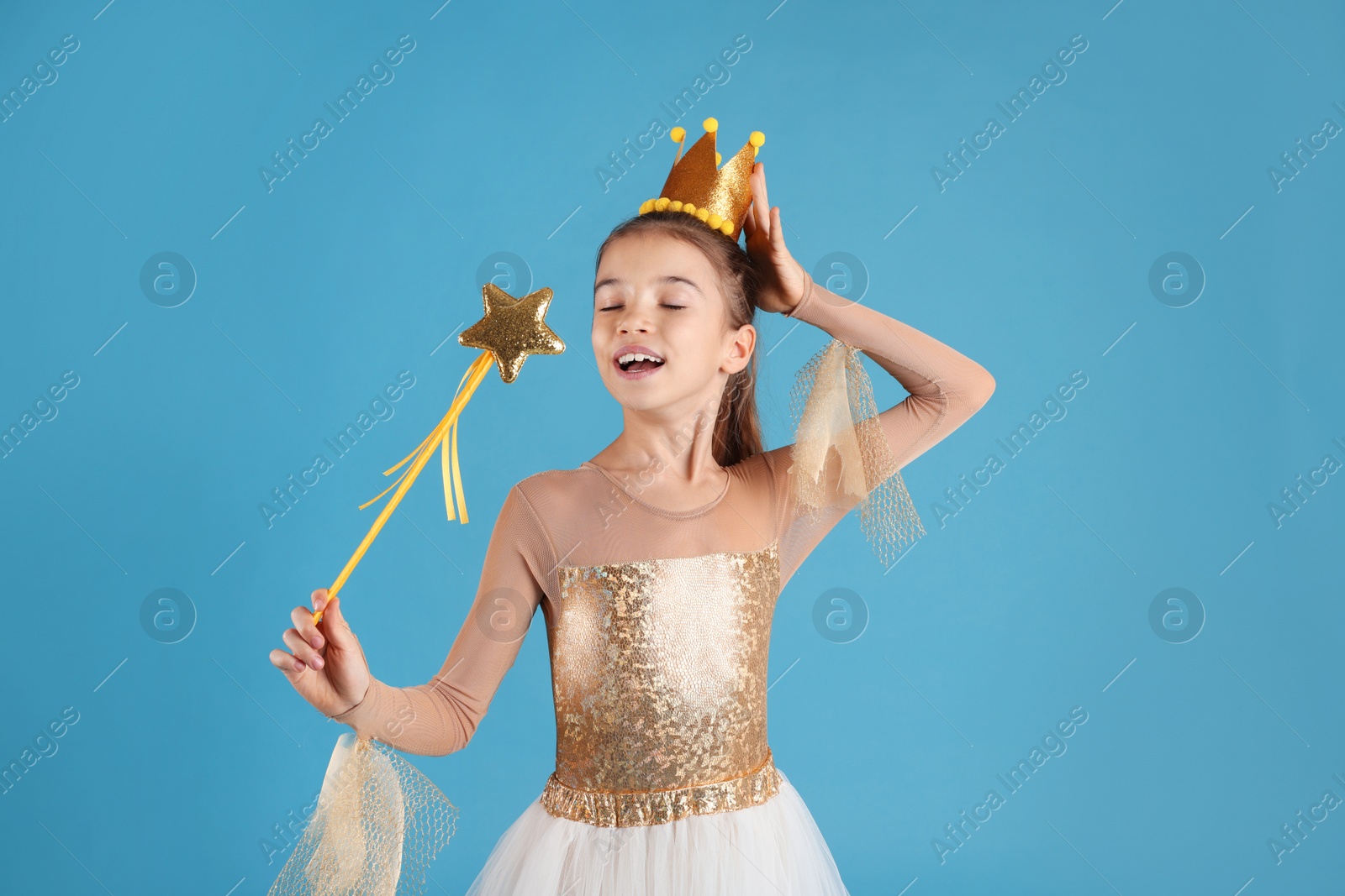 Photo of Cute girl in fairy dress with golden crown and magic wand on light blue background. Little princess