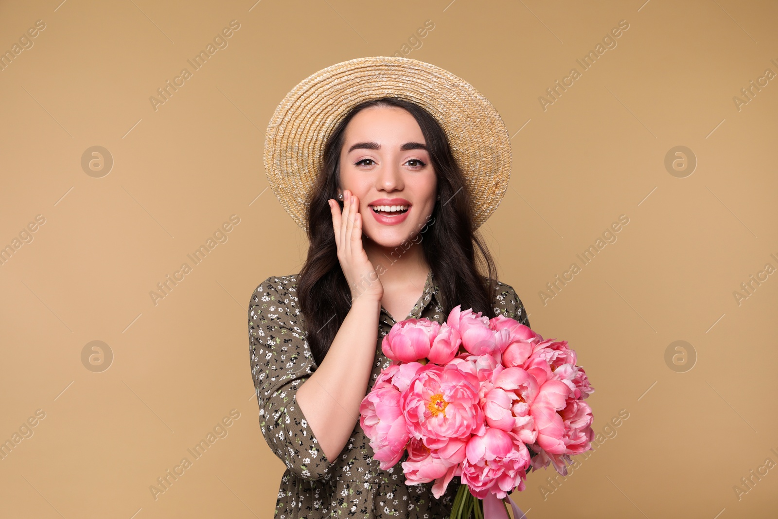 Photo of Beautiful young woman in straw hat with bouquet of pink peonies against light brown background