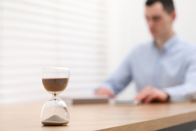 Hourglass with flowing sand on desk. Man using calculator indoors, selective focus