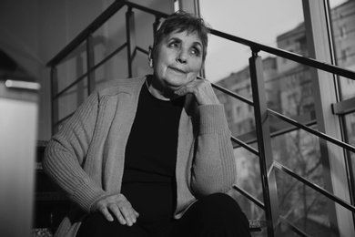 Photo of Poor senior woman sitting on stairs indoors. Black and white effect