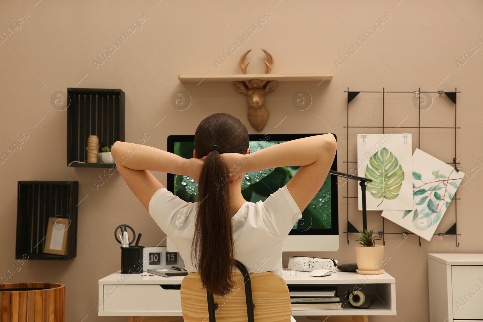 Photo of Woman sitting at white desk with computer near beige wall, back view. Interior design
