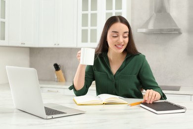 Photo of Home workplace. Happy woman opening notebook near laptop at marble desk in kitchen