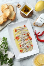 Photo of Tasty pickled anchovies with spices and products on white marble table, flat lay