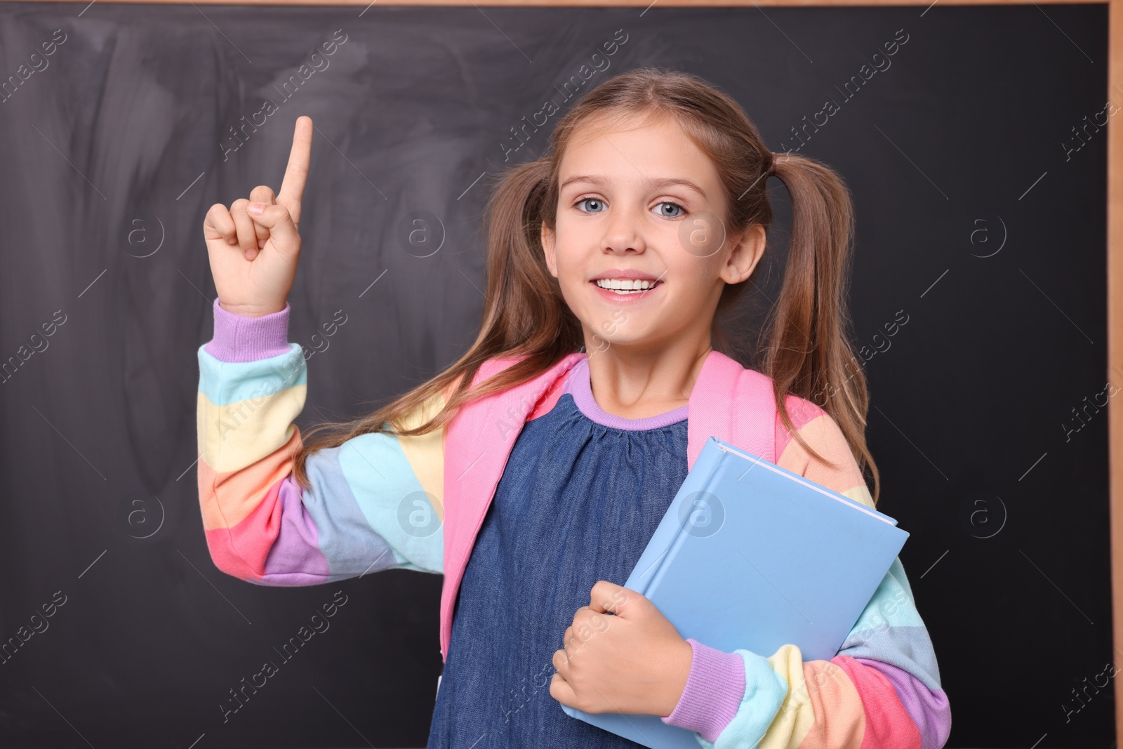 Photo of Smiling schoolgirl with book pointing at something near blackboard