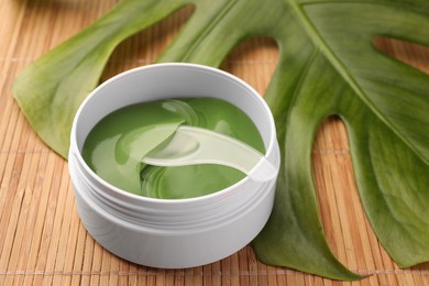 Photo of Jar of under eye patches with spoon and palm leaf on bamboo background, closeup. Cosmetic product