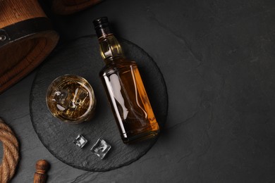 Whiskey with ice cubes in glass, bottle and barrel on black table, top view. Space for text