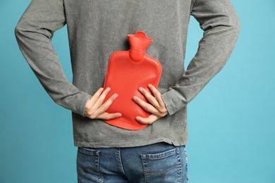 Photo of Man using hot water bottle to relieve back pain on light blue background, closeup