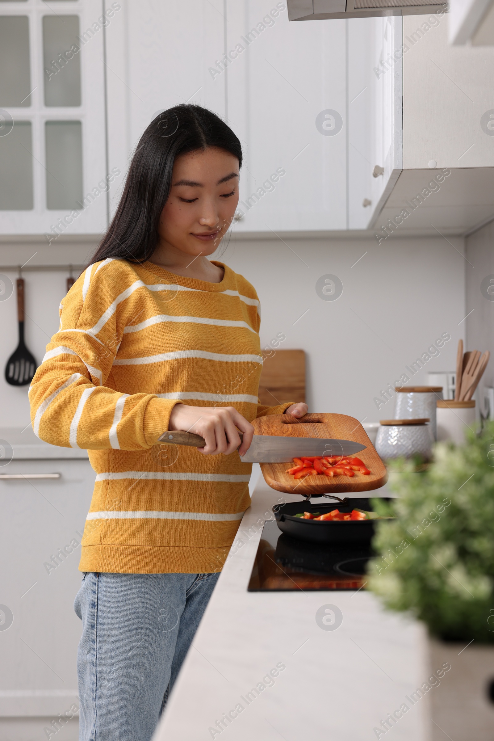 Photo of Cooking process. Beautiful woman adding cut bell pepper into pan with vegetables in kitchen