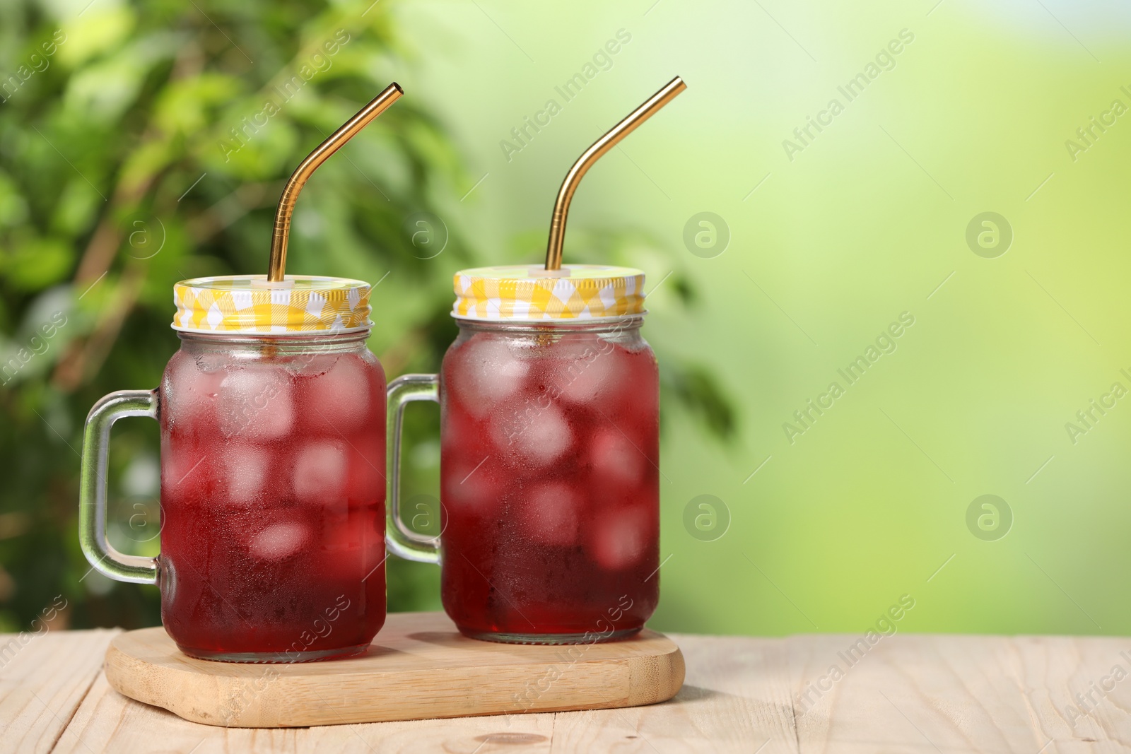 Photo of Refreshing hibiscus tea with ice cubes in mason jars on wooden table against blurred green background. Space for text