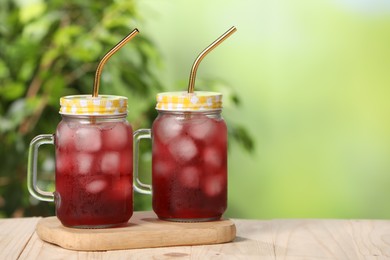 Photo of Refreshing hibiscus tea with ice cubes in mason jars on wooden table against blurred green background. Space for text