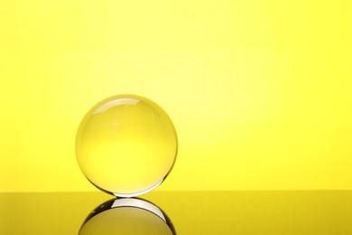 Photo of Transparent glass ball on mirror surface against yellow background. Space for text