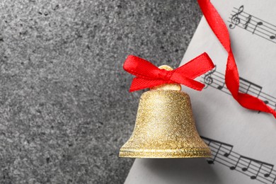 Photo of Golden shiny bell with red bow and music sheet on grey textured table, top view and space for text. Christmas decoration