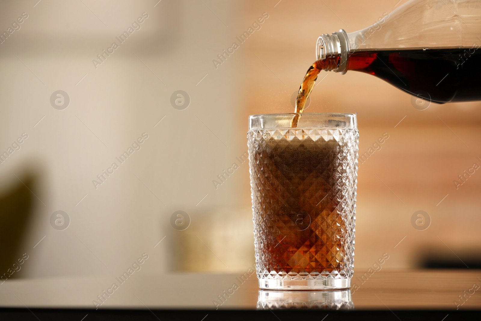 Photo of Pouring cola from bottle into glass with ice cubes on table against blurred background. Space for text