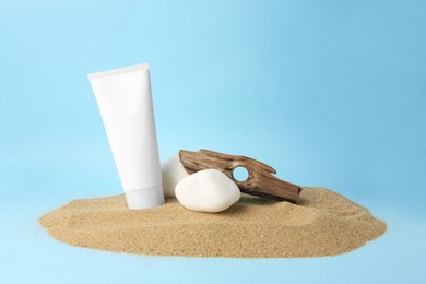 Photo of Tube of cream, stones and bark on sand against light blue background. Cosmetic product