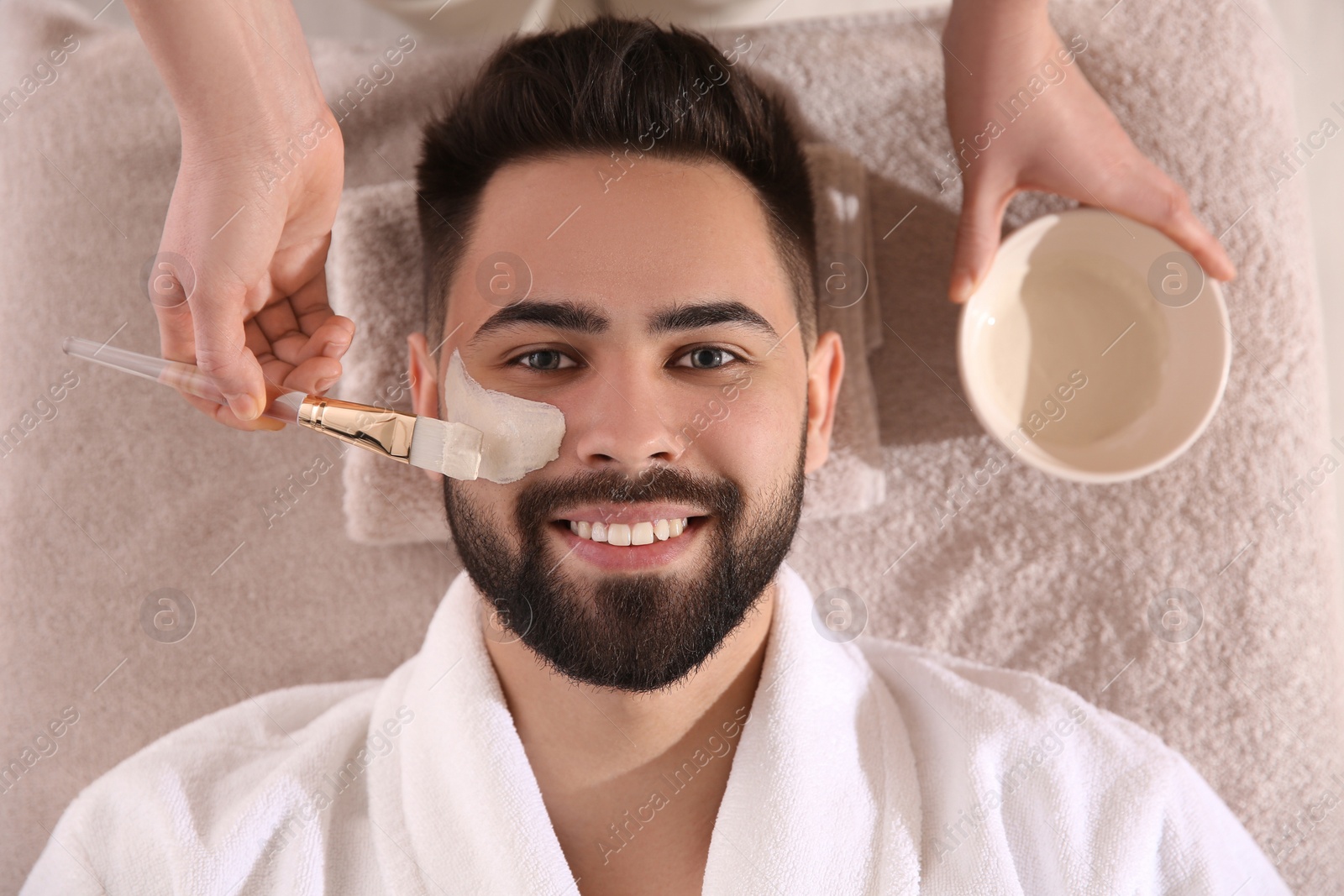 Photo of Cosmetologist applying mask on man's face in spa salon, top view