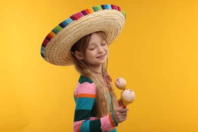 Photo of Cute girl in Mexican sombrero hat dancing with maracas on orange background