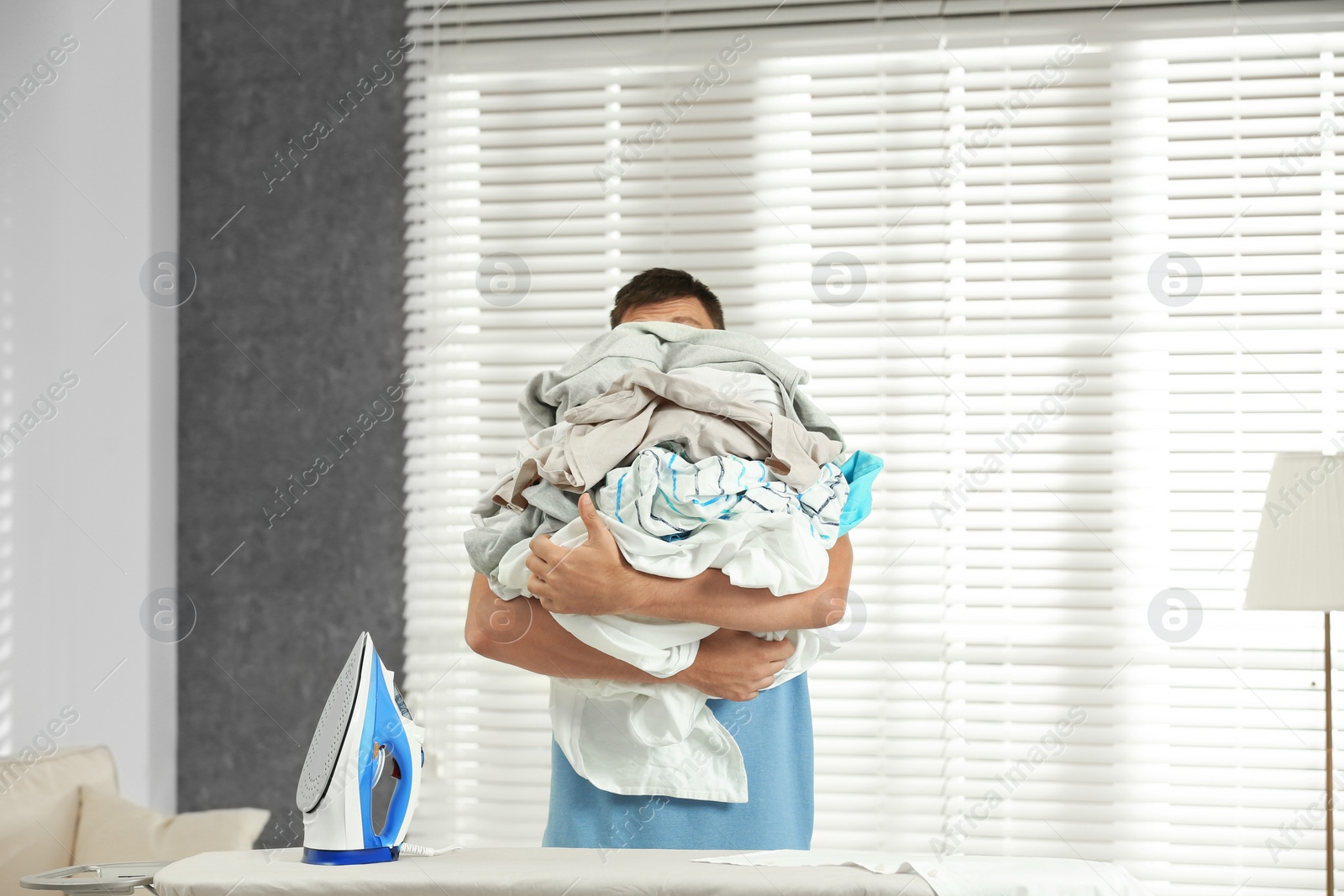 Photo of Handsome man with iron and unfolded laundry near board at home