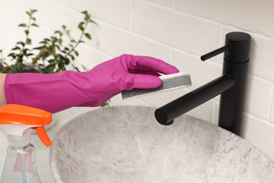 Photo of Woman in gloves cleaning faucet of bathroom sink with sponge, closeup