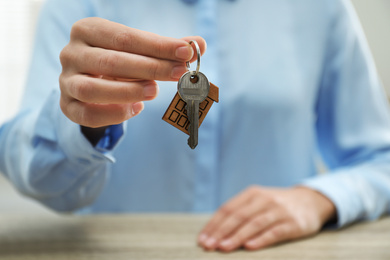 Photo of Real estate agent holding house key with trinket at wooden table, closeup