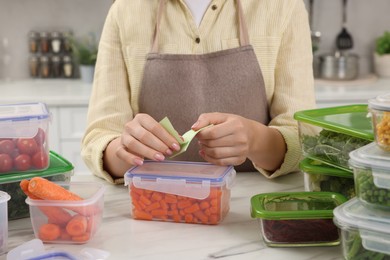 Photo of Woman sticking paper note onto container with fresh carrots at white marble table in kitchen, closeup. Food storage
