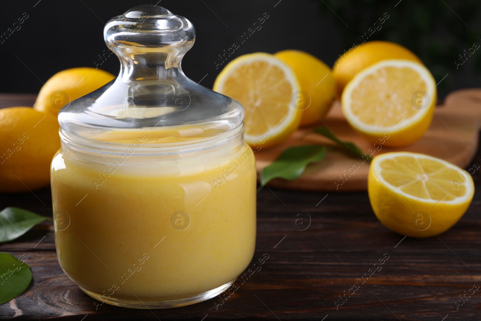 Photo of Delicious lemon curd in glass jar, fresh citrus fruits and green leaves on wooden table