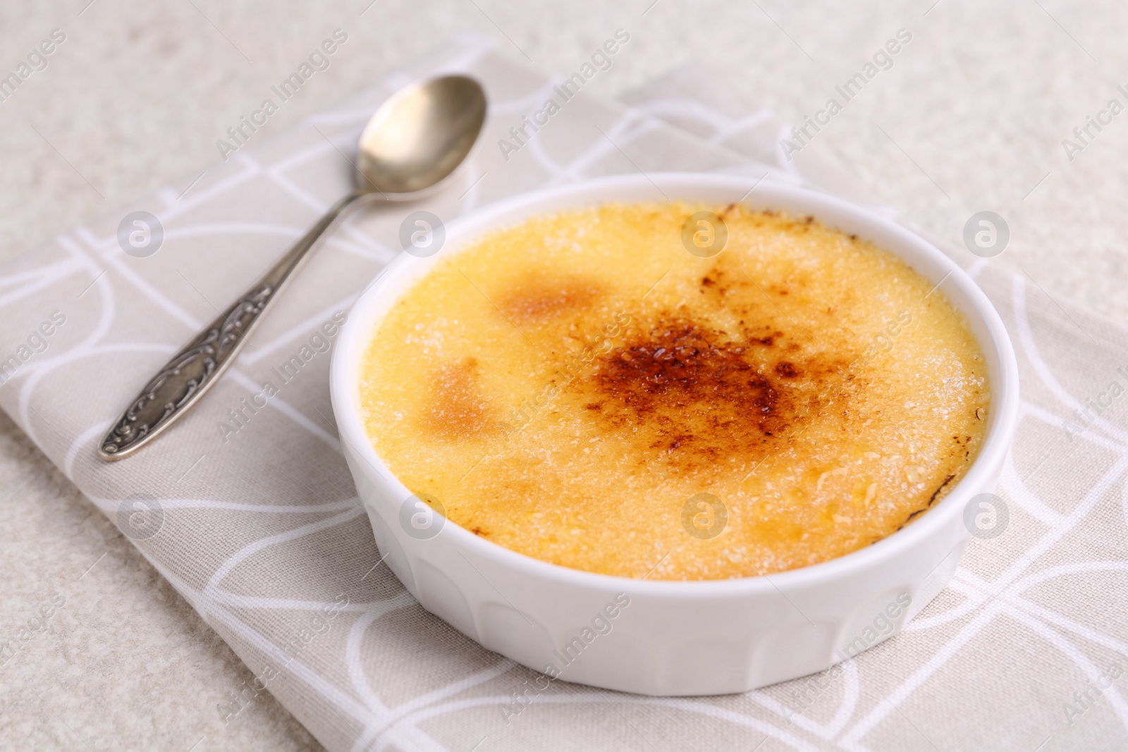 Photo of Delicious creme brulee in bowl and spoon on light textured table, closeup