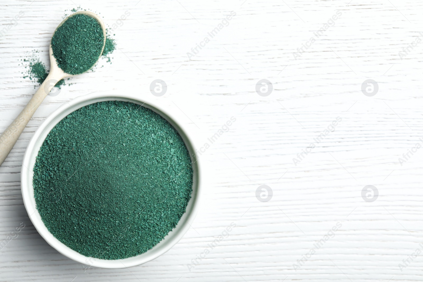 Photo of Bowl and spoon of spirulina powder on white wooden background, top view with space for text