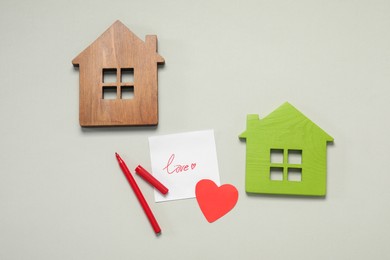 Long-distance relationship concept. Love note, paper heart and marker between two house models on light gray background, flat lay
