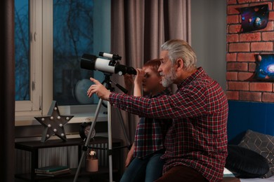 Photo of Little boy with his grandfather looking at stars through telescope in room