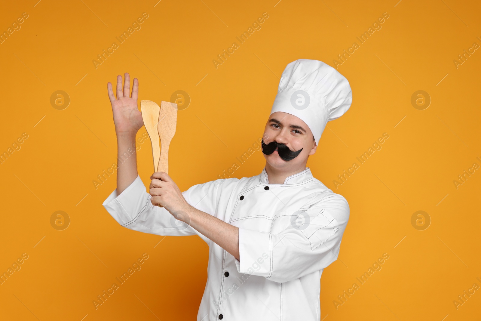 Photo of Portrait of happy confectioner with funny artificial moustache holding wooden spatulas on orange background