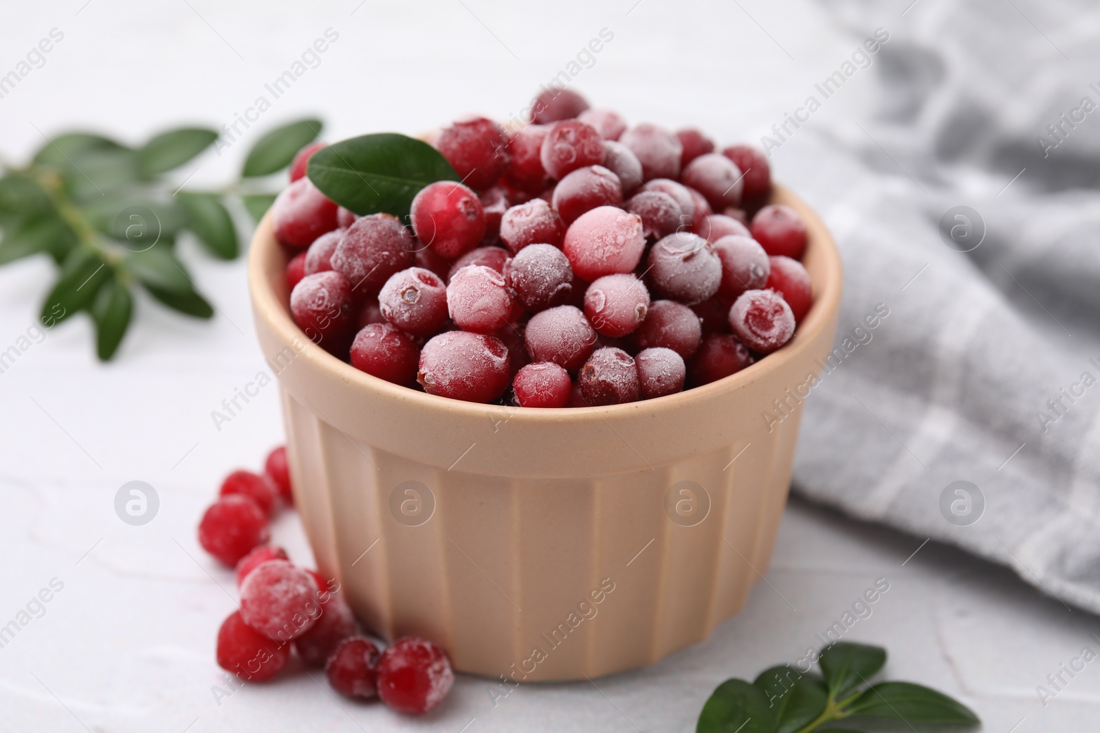 Photo of Frozen red cranberries in bowl and green leaves on white table, closeup