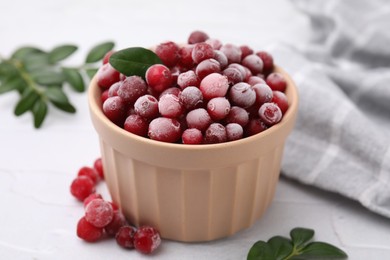 Frozen red cranberries in bowl and green leaves on white table, closeup