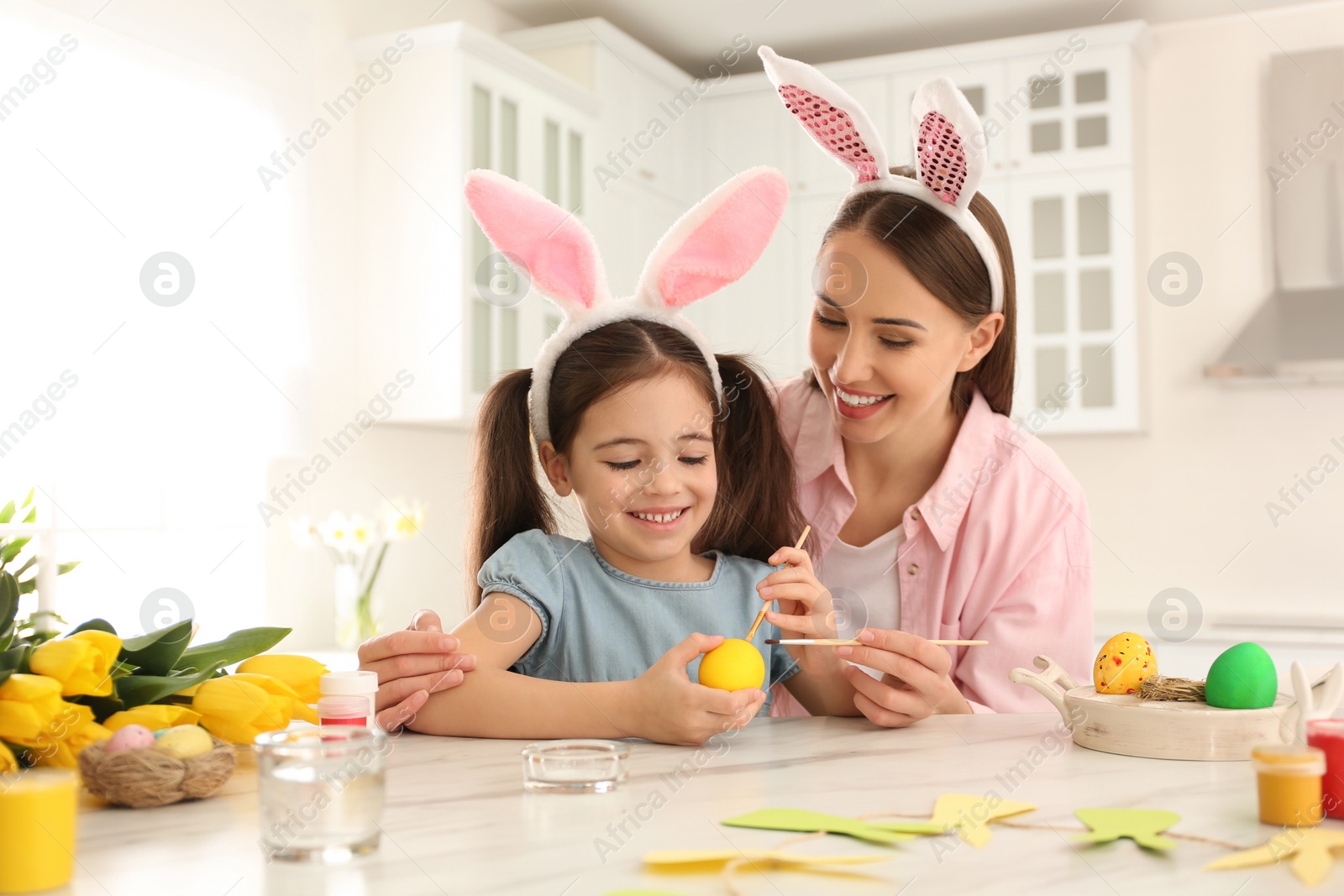 Photo of Happy mother and daughter with bunny ears headbands painting Easter egg in kitchen