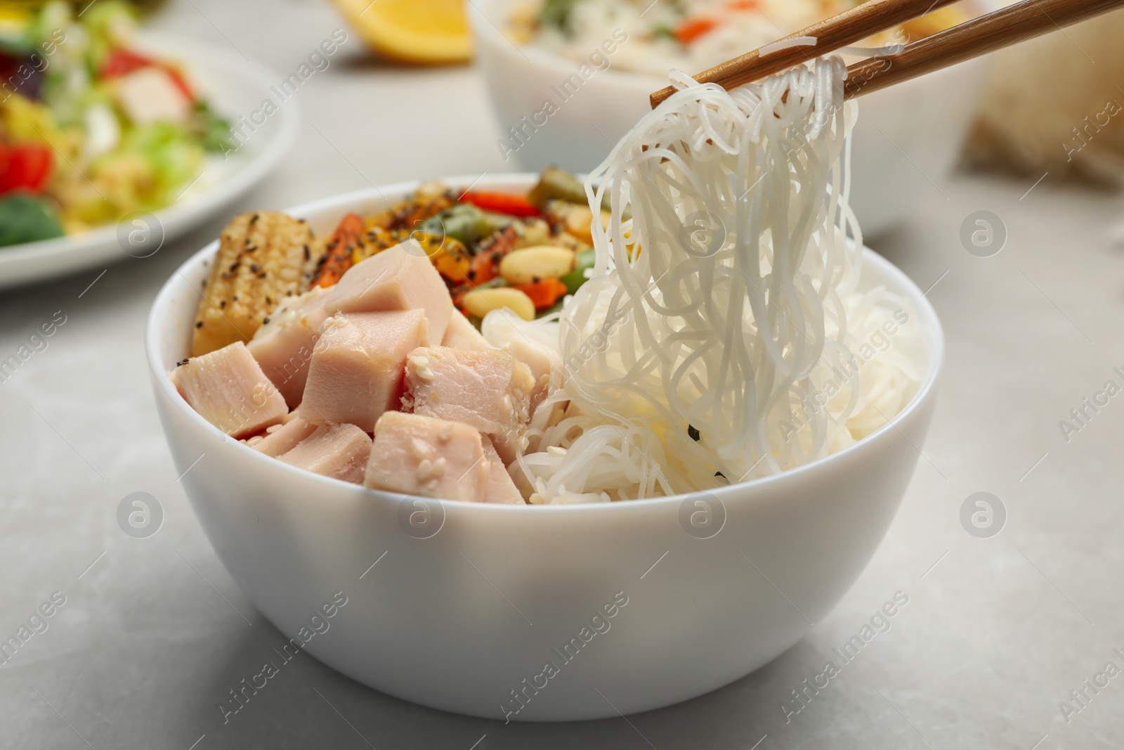 Photo of Chopsticks with tasty cooked rice noodles over bowl on grey table, closeup